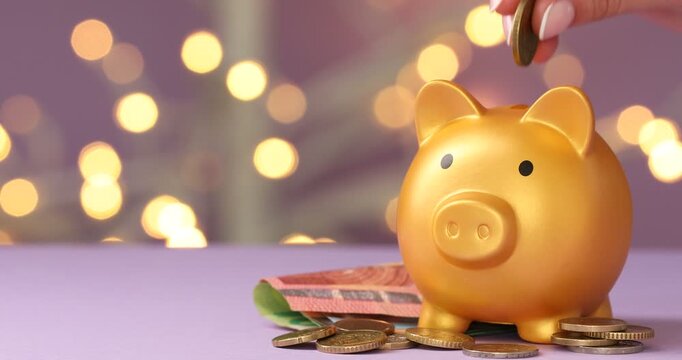 Woman putting coins into piggy bank against purple background with blurred lights, closeup. Bokeh effect