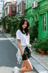 Fototapeta premium Young woman standing on residential street wearing white t shirt, black shorts, and sneakers, smiling with greenery and colorful houses behind her