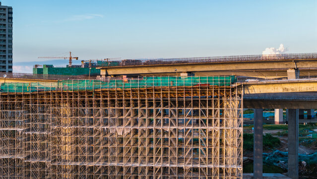 An urban construction site with dense scaffolding, an overpass under construction, and a distant building under a clear daytime sky.