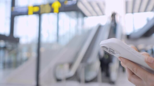 Hand holds phone, focuses on screen. Blurred airport background with escalators and travelers. Person seems calm, focused on device. Ideal for travel, communication, or technology themes