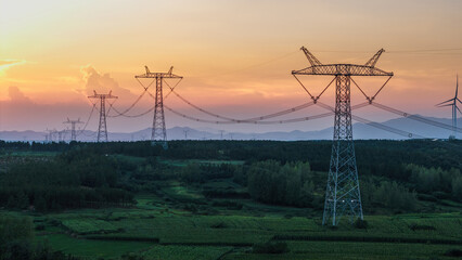 High-voltage power transmission towers extend across a green rural landscape at sunset, with wind turbines in the distance. © tai