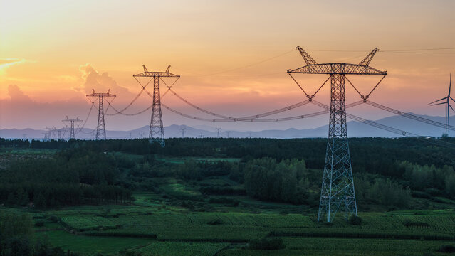 High-voltage power transmission towers extend across a green rural landscape at sunset, with wind turbines in the distance.
