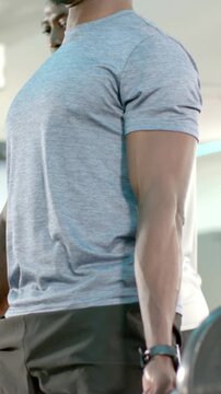 Vertical video: Bracing core, lifter in gray tee lifting loaded barbell in gym, spotter coaching