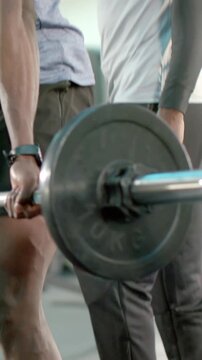 Vertical video: Positioning feet, man in gray tee gripping barbell in gym, pulling for strength