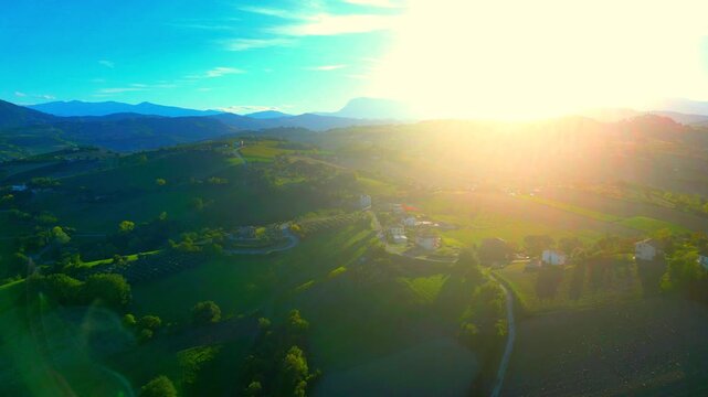 Impressive aerial pullback shot from Monte Rinaldo, prominent sunlight beaming down on the Marche hillscapes with gentle meadows, winding roads, fields, punctuated by trees, with Sibillini mountains