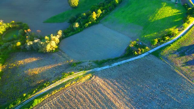 Suggestive aerial top-down bird's-eye tilt up view from Monte Rinaldo, from the countryside fields crossed by a gravel road to the nearby hills, trees, opening to a vision of stark Sibillini mountains