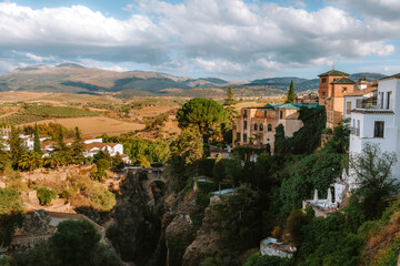 Obraz premium Rondas historic buildings perched atop the deep El Tajo gorge, revealing the ancient Puente Viejo bridge and surrounding Andalusian landscape with mountains under a cloudy sky