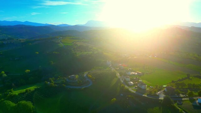 Enchanting aerial pullback shot from Monte Rinaldo, bright sunlight piercing through the Sibillini Mountains, reaching the hills, fields and meadows, imbuing them with spiritual qualities, blue skies