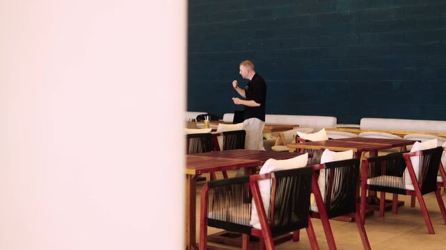 Man wearing casual clothes is standing in cafe and talking to someone gesturing his hands filmed in slow motion. Young adult male guy is engaged in business conversation in restaurant explaining topic