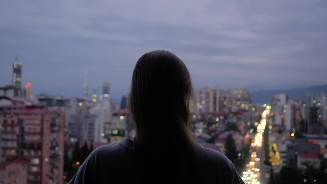 Back view: woman is standing on the balcony and looking at the evening sky over the city - central composition. Lonely, urban, dramatic and freedom concept