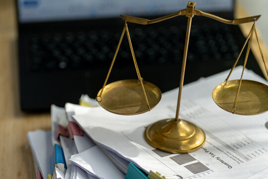 Golden scales sit atop a stack of papers. Files rest beside a laptop keyboard. Balance and law symbolize fair judgment. Use for legal, business, or finance concepts