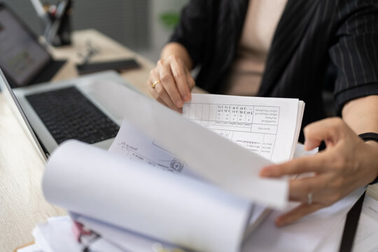 Woman reviews documents at desk with laptop nearby. Hands hold paper, fingers lightly tracing lines. Focused, thoughtful expression suggests concentration. Ideal for business, education