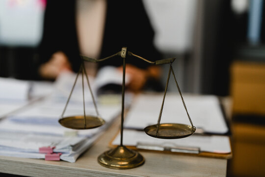 Golden scales sit on a wooden desk. Blurry papers and folders surround the symbol. Person in dark clothes stands nearby. The scene suggests legal or judicial matters. Suitable for law, justice
