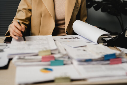 Woman sits at desk, surrounded by paperwork. She holds pen, reviewing documents with focus. Stacks of papers, sticky notes, charts clutter the table