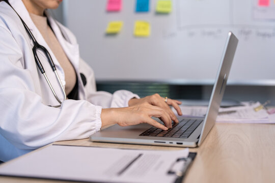 Doctor types on laptop at desk. Stethoscope rests around her neck. Whiteboard with sticky notes behind her. Focused, calm, professional demeanor. Ideal for healthcare or medical tech scenes