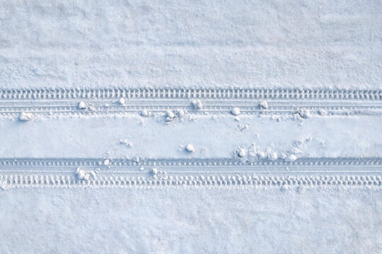 Top-down view of two parallel tire tracks imprinted on fresh, undisturbed white snow, with small snow clumps scattered along the tracks.
