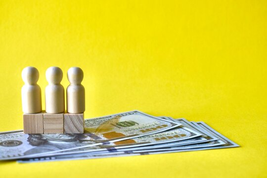 Three identical white wooden figures standing on wooden blocks on a row of one-hundred-dollar bills, representing team salary, corporate payroll, and equal wage standards.