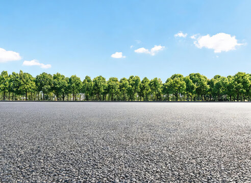 A smooth asphalt road stretches forward, flanked by dense green trees under a clear blue sky with scattered white clouds, presenting a serene outdoor landscape.