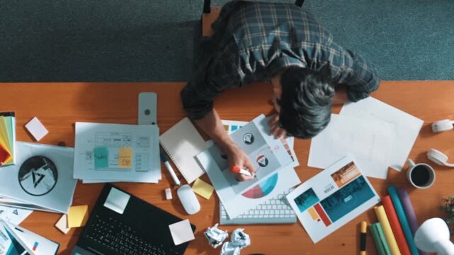 Top view of designer writing and designing logo graphic at meeting. Closeup of business man working on colored palettes and writing idea on sticky notes on table with equipment and paper. Symposium.