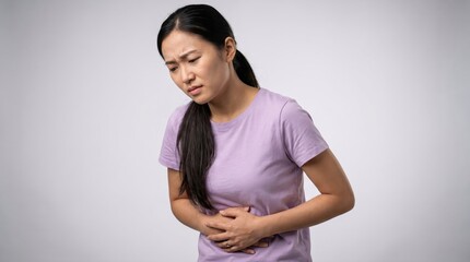 Obraz premium Young asian woman wearing a light purple t shirt holding her stomach in pain with a distressed facial expression against a plain light gray studio background