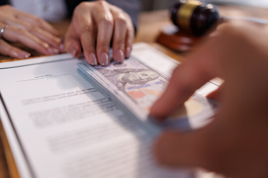 Hand passing stack of hundred dollar banknotes over contract document, symbolizing acts of bribery, corruption, illegal payment, and judicial wrongdoing in legal proceedings