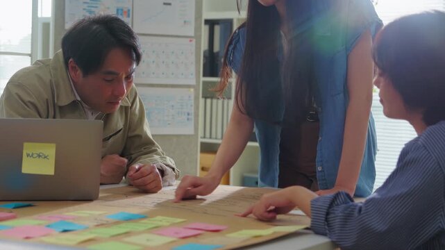 Three colleagues lean over a desk examining a hand-drawn business strategy map covered in colorful sticky notes, brainstorming project ideas together beside a laptop in a bright modern office.