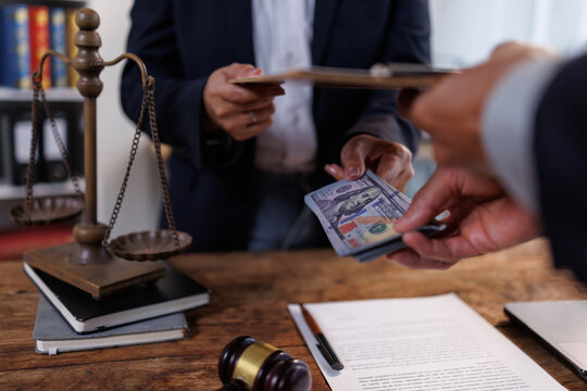 Hands of individuals exchanging a stack of us dollar currency notes, symbolizing bribery and corruption in legal or financial contexts, with scales of justice and a gavel nearby