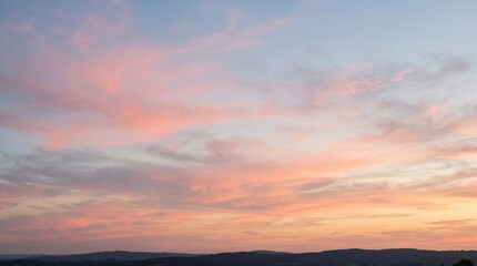 Fototapeta premium Soft pink and orange sunset clouds stretching across a pale blue sky above a dark silhouetted horizon line of rolling hills at dusk