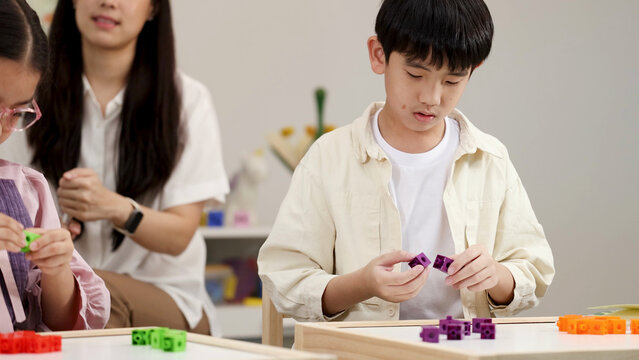 Asian Boy Focusing on Math Manipulative Blocks with Teacher in Background