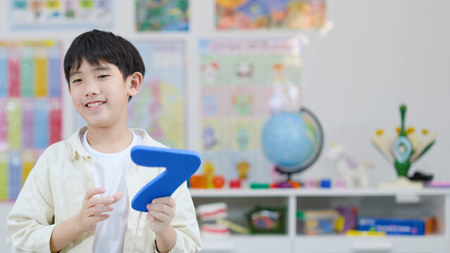 Smiling Asian Schoolboy Holding Blue Foam Number Seven in Classroom