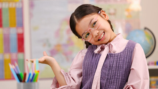 Cheerful Asian Schoolgirl in Pink Glasses Gesturing in Classroom