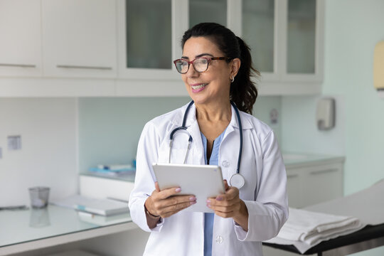 Happy senior practitioner doctor woman in glasses and white coat holding tablet in medical professional office, looking away, smiling, thinking on online communication with patient