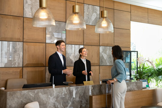 Female guest standing at hotel reception desk with suitcase during check-in, talking with receptionists, confirming reservation details. Hospitality, customer services concept for travel and tourism