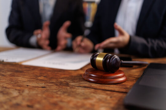 Gavel on a rustic wooden desk, two business professionals discussing legal documents during a consultation, symbolizing justice, arbitration, and agreement in a professional setting