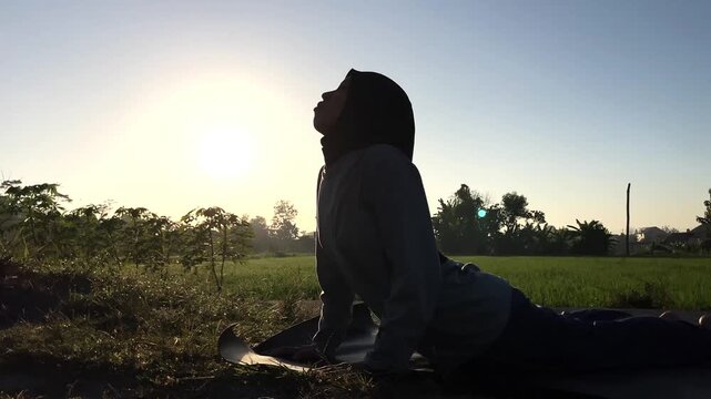 Slow motion clip of a Muslim woman in a hijab doing cobra yoga pose on a mat outdoors. Beautiful golden sunrise backlight over a green field. Perfect for wellness and fitness concepts.