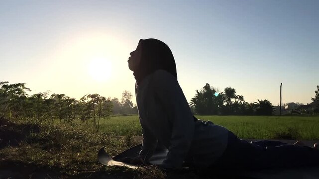 Slow motion clip of a Muslim woman in a hijab doing cobra yoga pose on a mat outdoors. Beautiful golden sunrise backlight over a green field. Perfect for wellness and fitness concepts.
