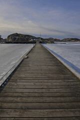 Fototapeta premium Symmetrical view of a wooden pier with rope railings leading across the ice in Skrhamn. A cold winter day in the Swedish archipelago with dramatic light and frozen wate