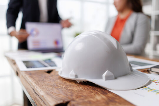 White hard hat lying on a rustic wooden table, while construction and engineering professionals collaborate, examining project plans and financial data during a strategic work meeting
