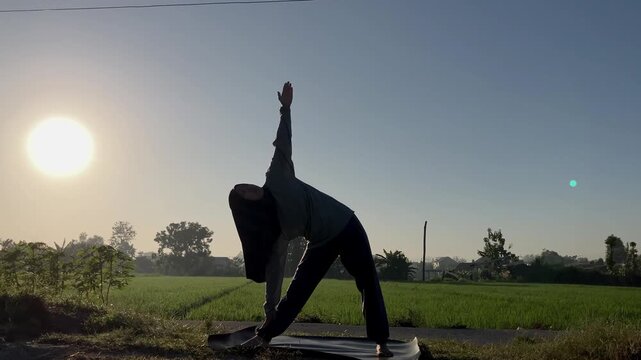 Slow motion silhouette of a Muslim woman in a hijab doing the triangle yoga pose (Trikonasana) on a mat outdoors. Beautiful golden sunrise over a lush green rice field. Perfect for wellness themes.
