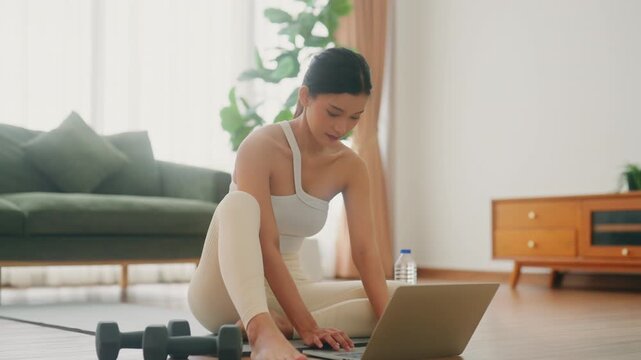 Young Asian woman sitting on the floor on yoga mat in front of laptop and training at home. Beautiful smiling woman doing stretching exercise while communicates with a trainer online before workout