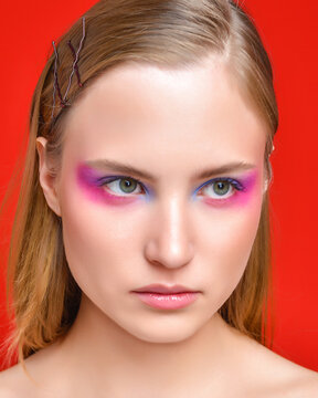 A woman with bright, expressive eye makeup on a red background. Her expression is serious, focused, and confident.