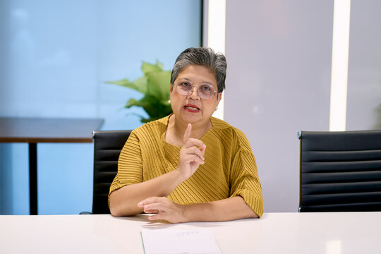 An experienced senior Asian businesswoman in a yellow top and glasses is speaking and gesturing with her finger during a presentation or meeting. She sits at a white conference table in a modern, brig