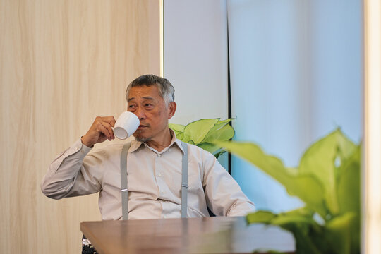 Senior Asian Man with Suspenders Drinking Coffee in Modern Indoor Setting, Thoughtful Elderly Male Professional Relaxing at Cafe or Office Table.