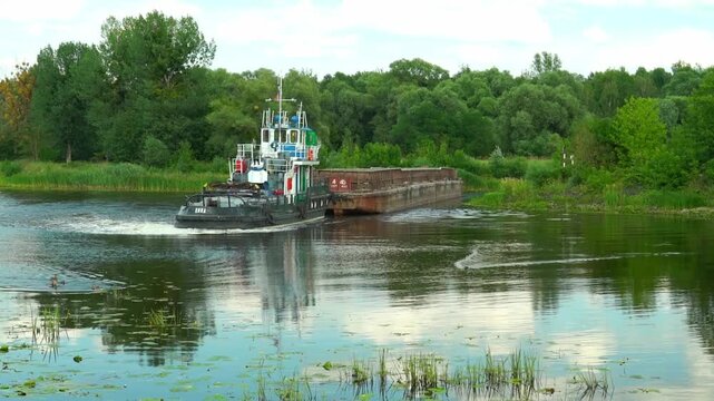 A tugboat pulls a large barge along the waterway in a lush green area. This shows the vessel's steady movement through the calm waters under a blue sky.in Belarus, in the city of Pinsk, on the embankm