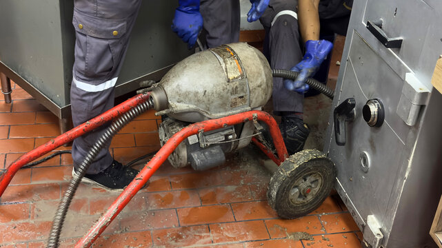Professional plumbers operating an electric drain cleaner machine, feeding a flexible steel auger cable to clear a clogged pipe on a commercial kitchen floor.