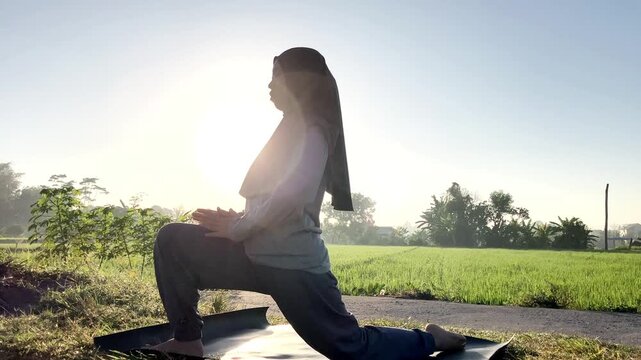 Slow motion side view of a Muslim woman in a hijab doing a low lunge yoga pose on a mat outdoors. Beautiful bright sunrise backlight over a green rice field. Perfect for wellness and health.