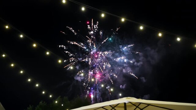 Colorful fireworks illuminate the night sky above an outdoor venue decorated with string lights. Festive pyrotechnic display creates a vibrant atmosphere during an evening celebration.