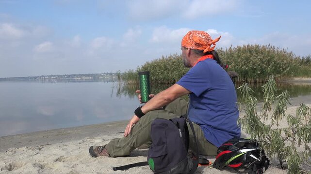 Traveler relaxing by a peaceful lake, drinking from a reusable travel mug during a bicycle touring break on a sandy lakeshore surrounded by nature