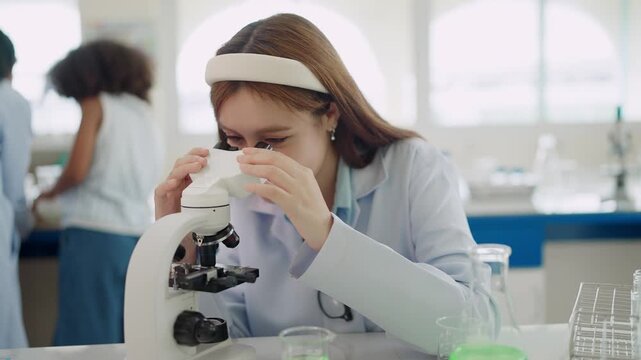 Young scientist smiles while examining samples through microscope. Hands adjust focus knob with care and precision. Bright lab holds test tubes and glassware in background. Scene suggests curiosity