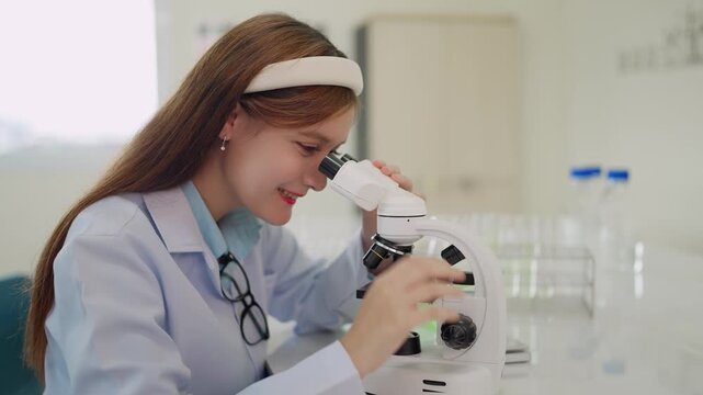 Young scientist smiles while examining samples through microscope. Hands adjust focus knob with care and precision. Bright lab holds test tubes and glassware in background. Scene suggests curiosity